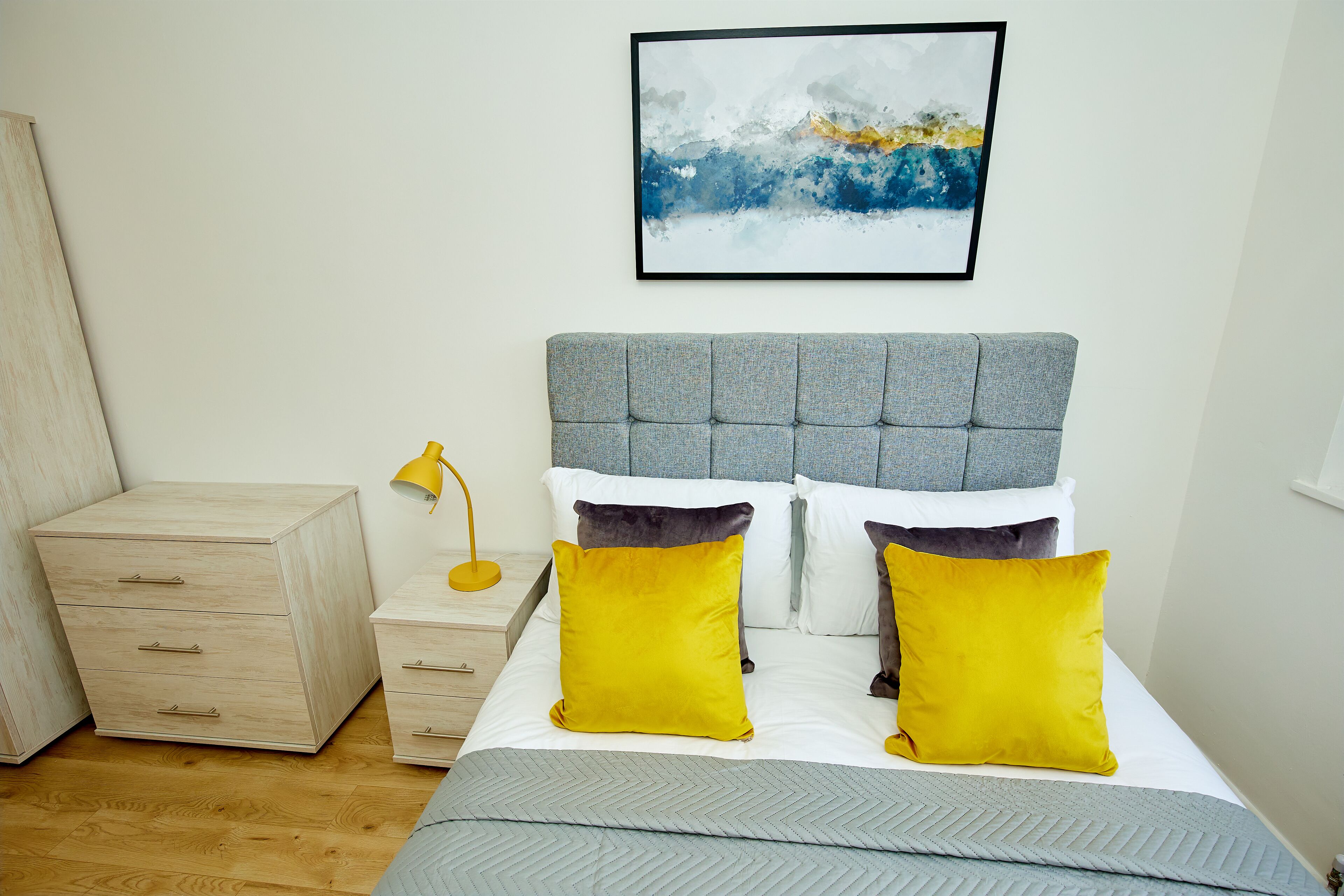 A bedroom with a bed, a nightstand, a drawer unit, and a wall-mounted abstract painting.