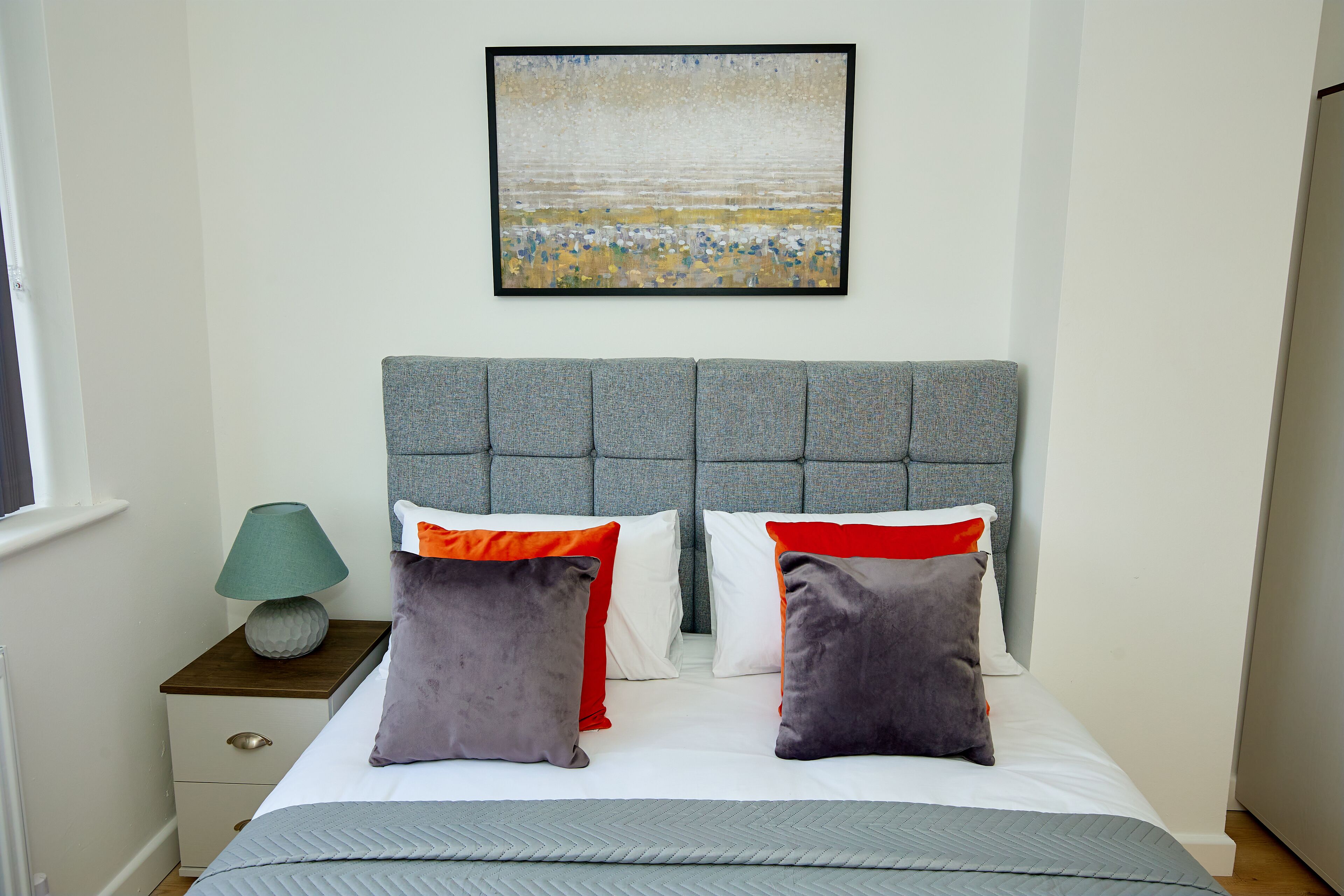 A neatly made bed with grey bedding, a grey headboard, and two grey pillows with red and orange accents.