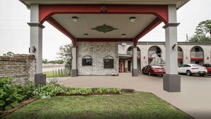 A building with a brick facade, a covered entrance area with a red roof, and a parking lot with cars. - Hollywood Inn (Houston)