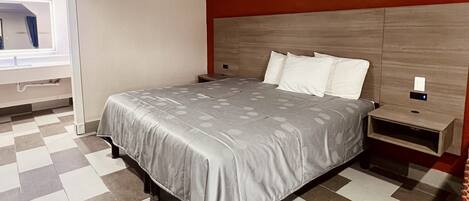 A hotel room with a checkered floor, a bed with a grey bedspread, a wooden headboard, a nightstand, and a mirror with a light above the sink.