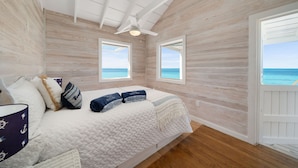Une chambre avec un plafond en bois, un lit recouvert d’une couette blanche, deux fenêtres donnant sur l’océan et une porte menant à l’extérieur.