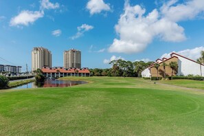 A golf course with a pond, tall buildings, and a white building with a red roof. - Beachfront Days Ocean View Condo Bliss (Panama City Beach)