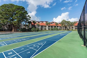 A shuffleboard court with a clear view of a building with red roofs and palm trees in the background. - Beachfront Days Ocean View Condo Bliss (Panama City Beach)