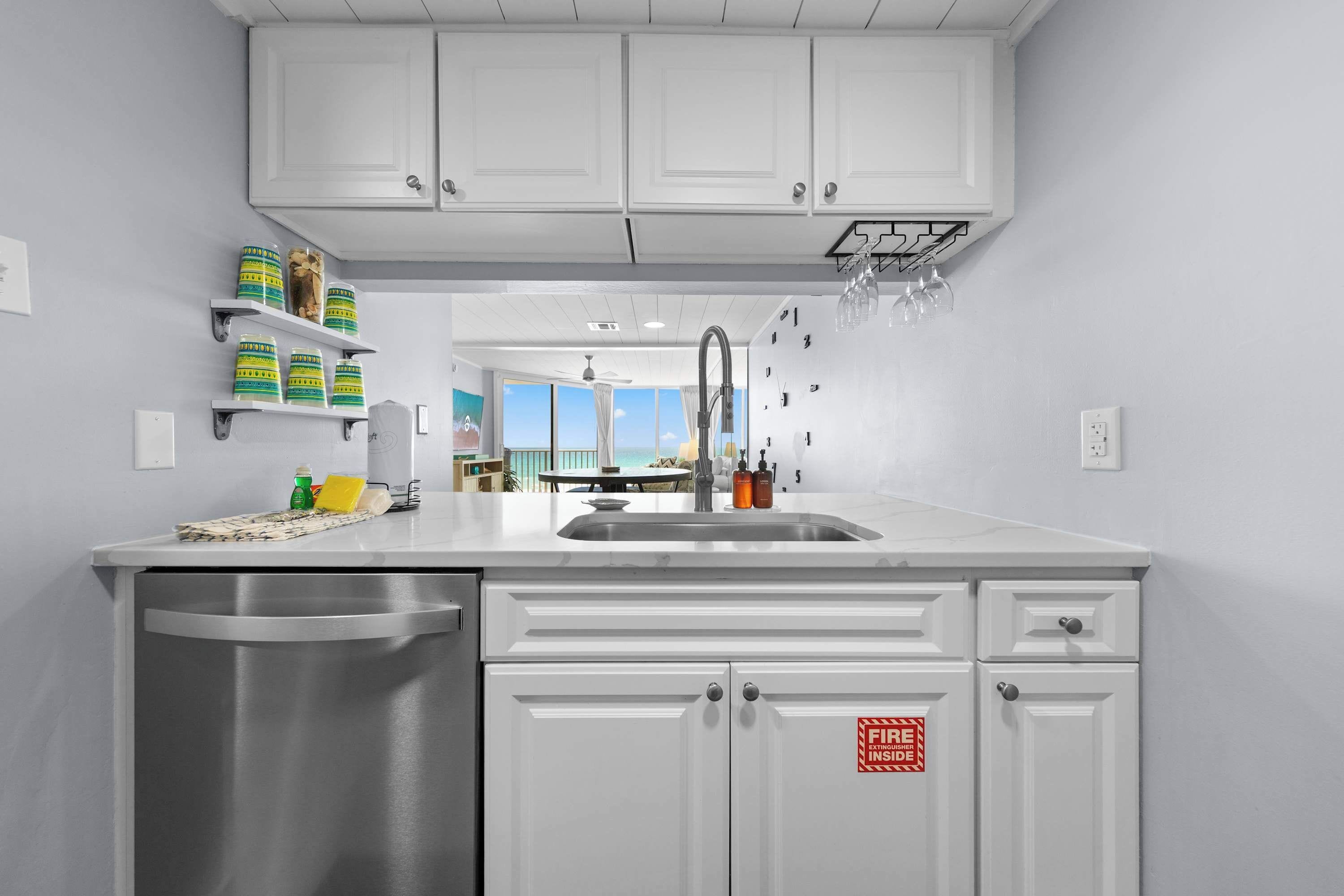 A modern kitchen with white cabinets, a stainless steel dishwasher, and a sink. There are shelves with jars and a view of the ocean through large windows.