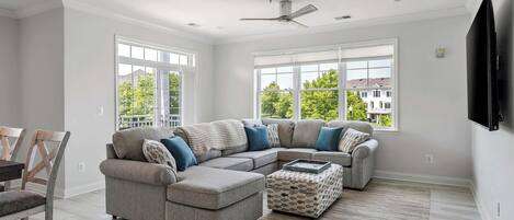 A spacious living room with a sectional sofa, ottoman, and a flat-screen TV mounted on the wall.