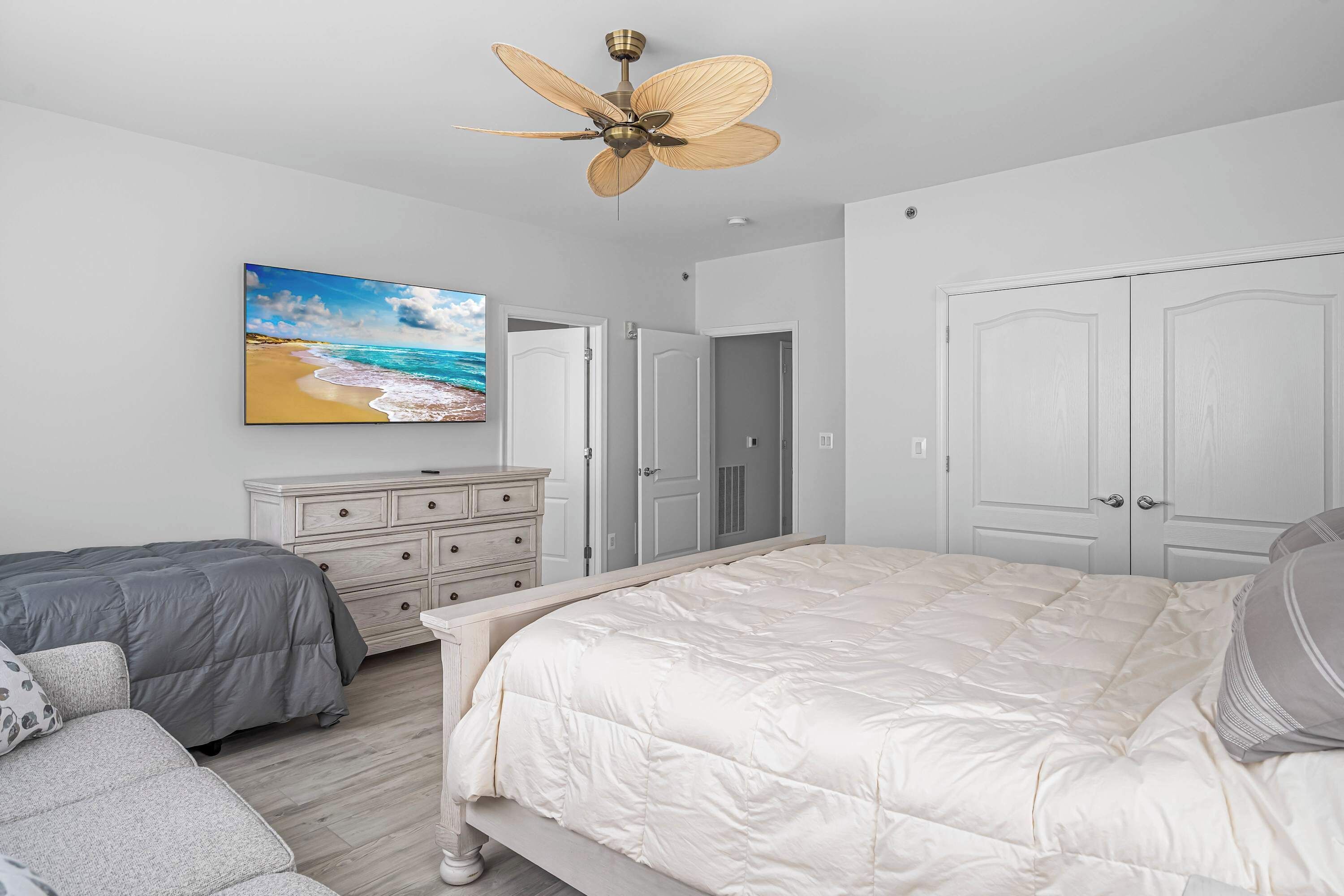 A bedroom with a large bed, a ceiling fan, a dresser, and a painting of a beach scene.