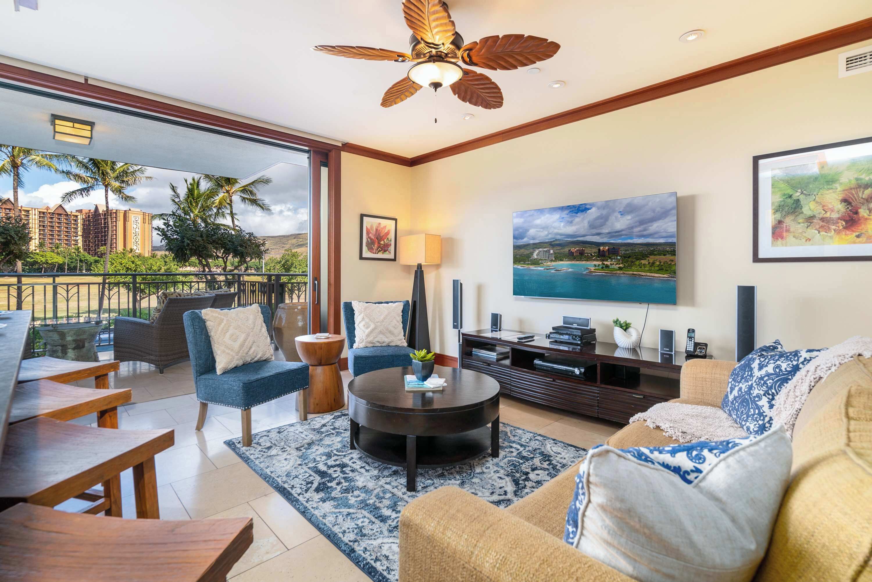 A spacious living room with a large TV, comfortable seating, a ceiling fan, and a view of the cityscape through large windows.