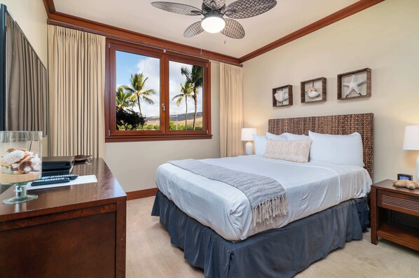 Habitación de hotel con una cama grande, un escritorio con una computadora y un vaso de postre, un ventilador de techo y vista a palmeras a través de una ventana.
