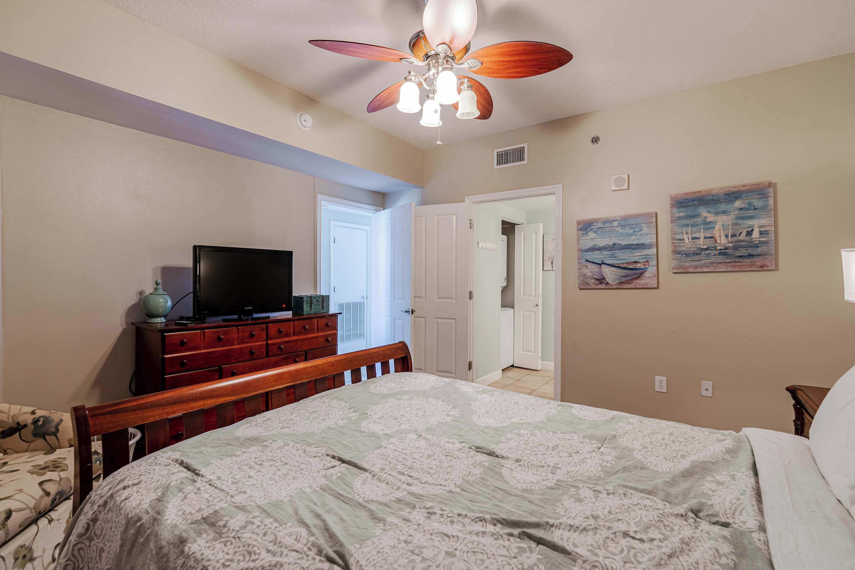 A bedroom with a bed, wooden dresser, television, ceiling fan, and two paintings on the wall.