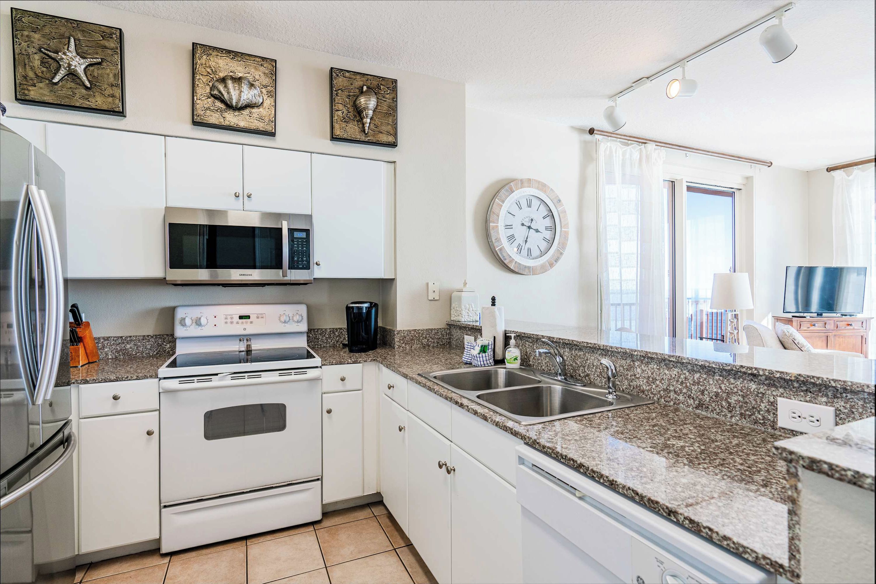 A modern kitchen with white cabinets, a stainless steel refrigerator, a microwave, an oven, and a granite countertop. There are decorative shells on the wall and a clock.