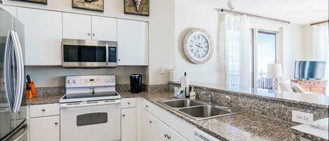 A modern kitchen with white cabinets, a stainless steel refrigerator, a microwave, an oven, and a granite countertop. There are decorative shells on the wall and a clock.