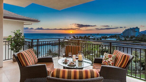 A balcony with wicker furniture, a glass table, and a sunset view. - Hale Leilani Ko Olina Beachfront w/ Views! (Kapolei)