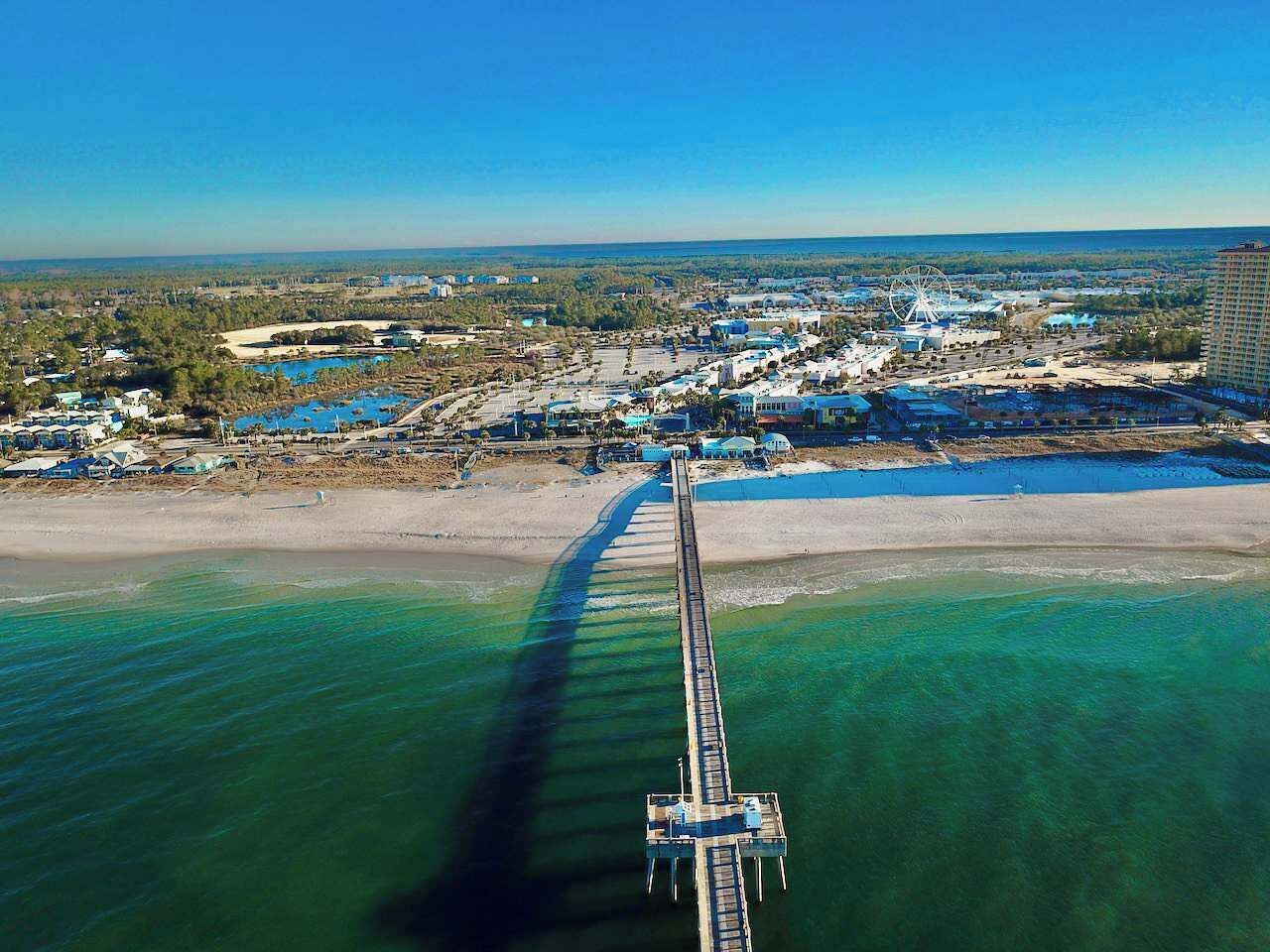 An aerial view of a coastal area with a long pier extending into the water, surrounded by buildings and a clear sky.