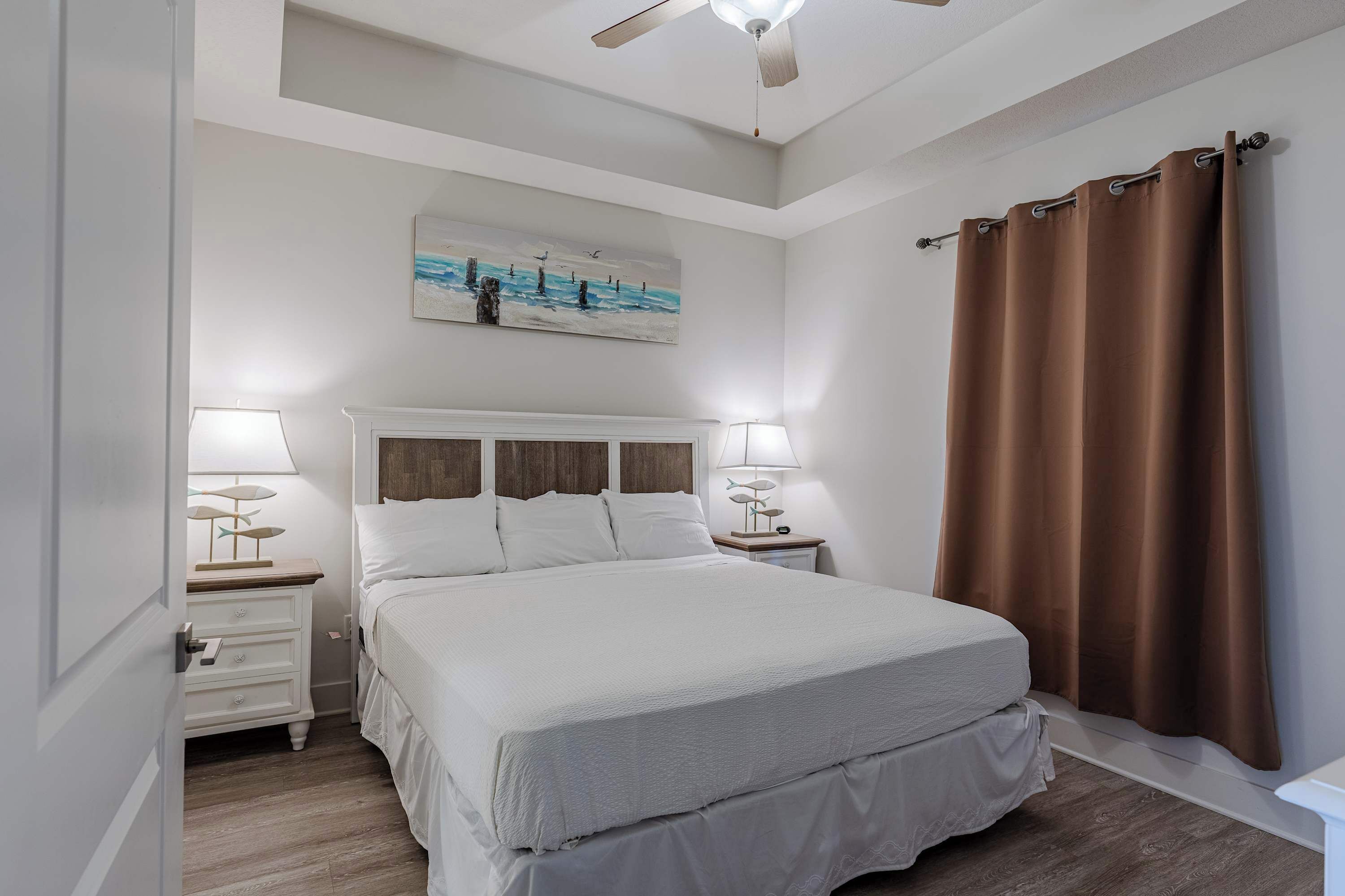 A bedroom with a large bed, two bedside tables with lamps, a ceiling fan, a painting on the wall, and brown curtains.