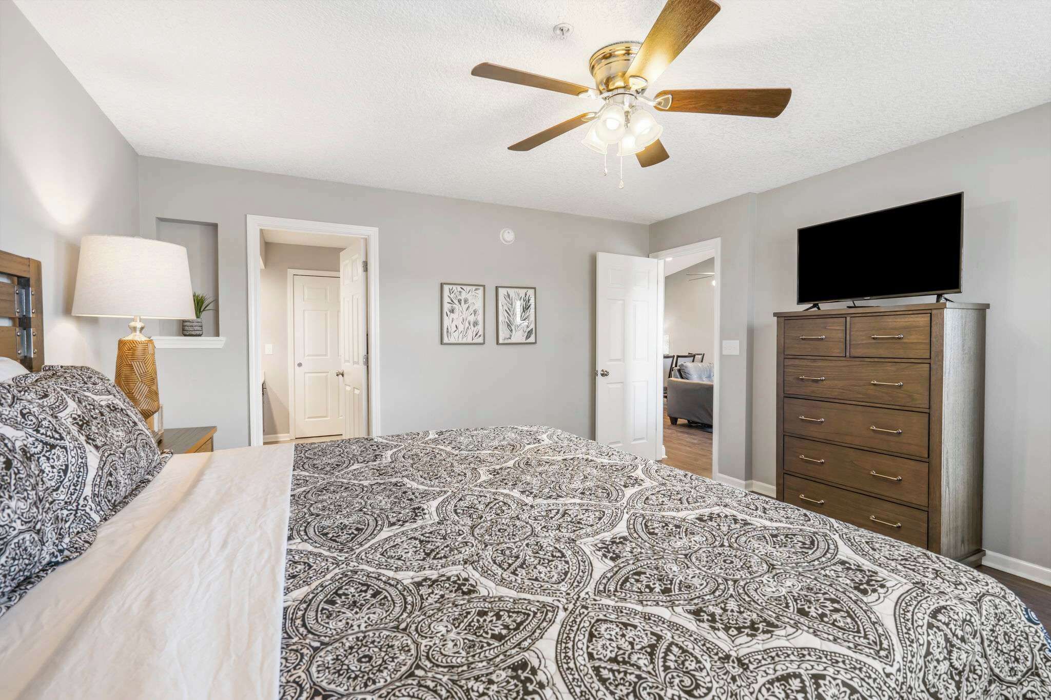 Une chambre avec un grand lit, un ventilateur de plafond, un téléviseur posé sur une commode et une porte donnant sur une autre pièce.