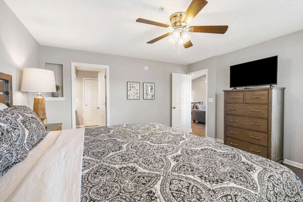 Une chambre avec un grand lit, un ventilateur de plafond, un téléviseur posé sur une commode et une porte donnant sur une autre pièce.