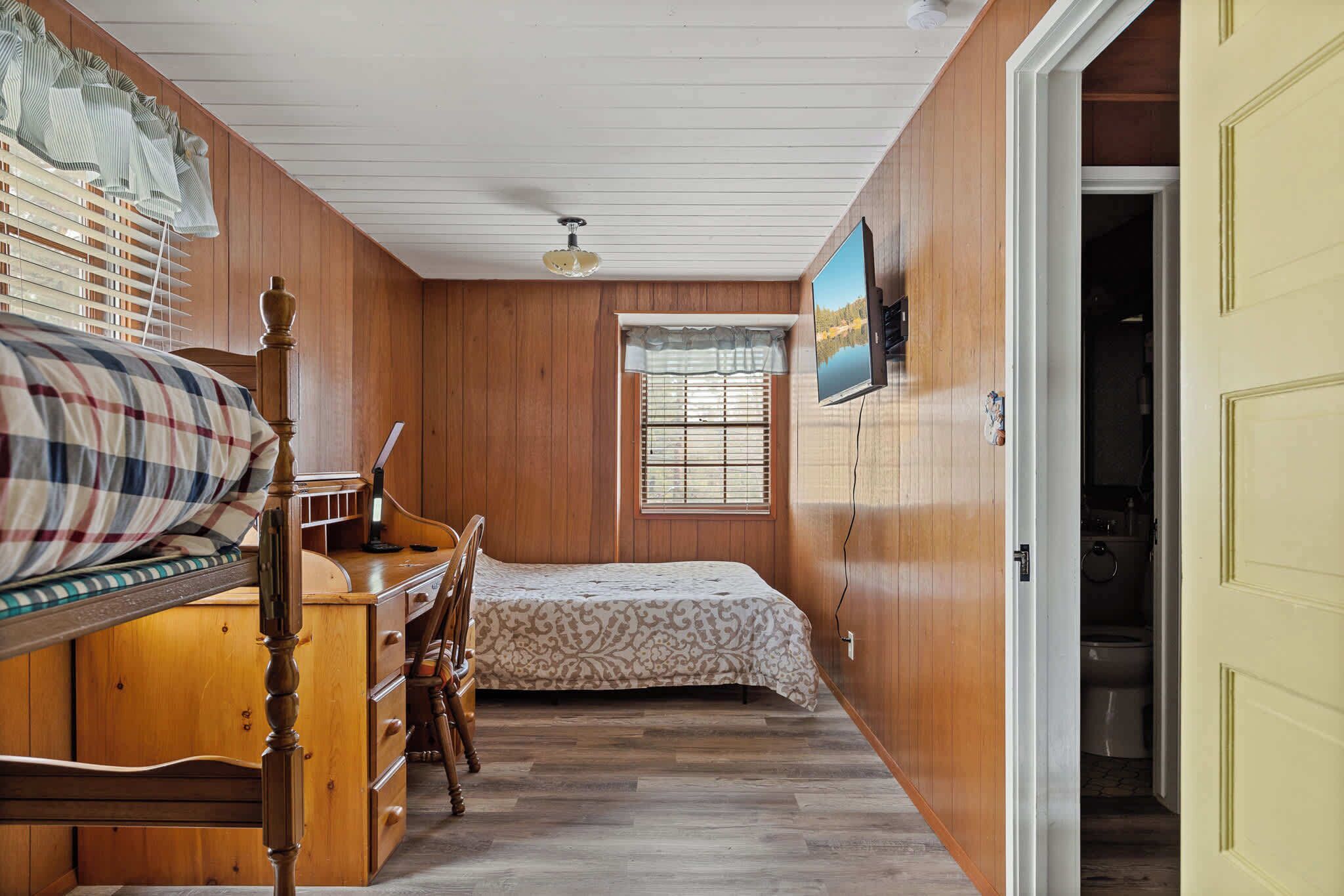 Un dormitorio con una cama de madera, un tocador, una silla, una ventana con persianas y un televisor en la pared.