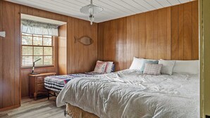 Ein Schlafzimmer mit Holzwänden, einem Bett mit vielen Kissen, einem Schreibtisch mit Lampe und einem Fenster mit Jalousien.