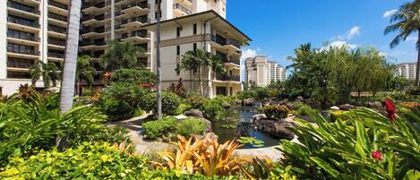 A hotel with multiple floors, a garden with various plants, a small pond, and a clear blue sky.