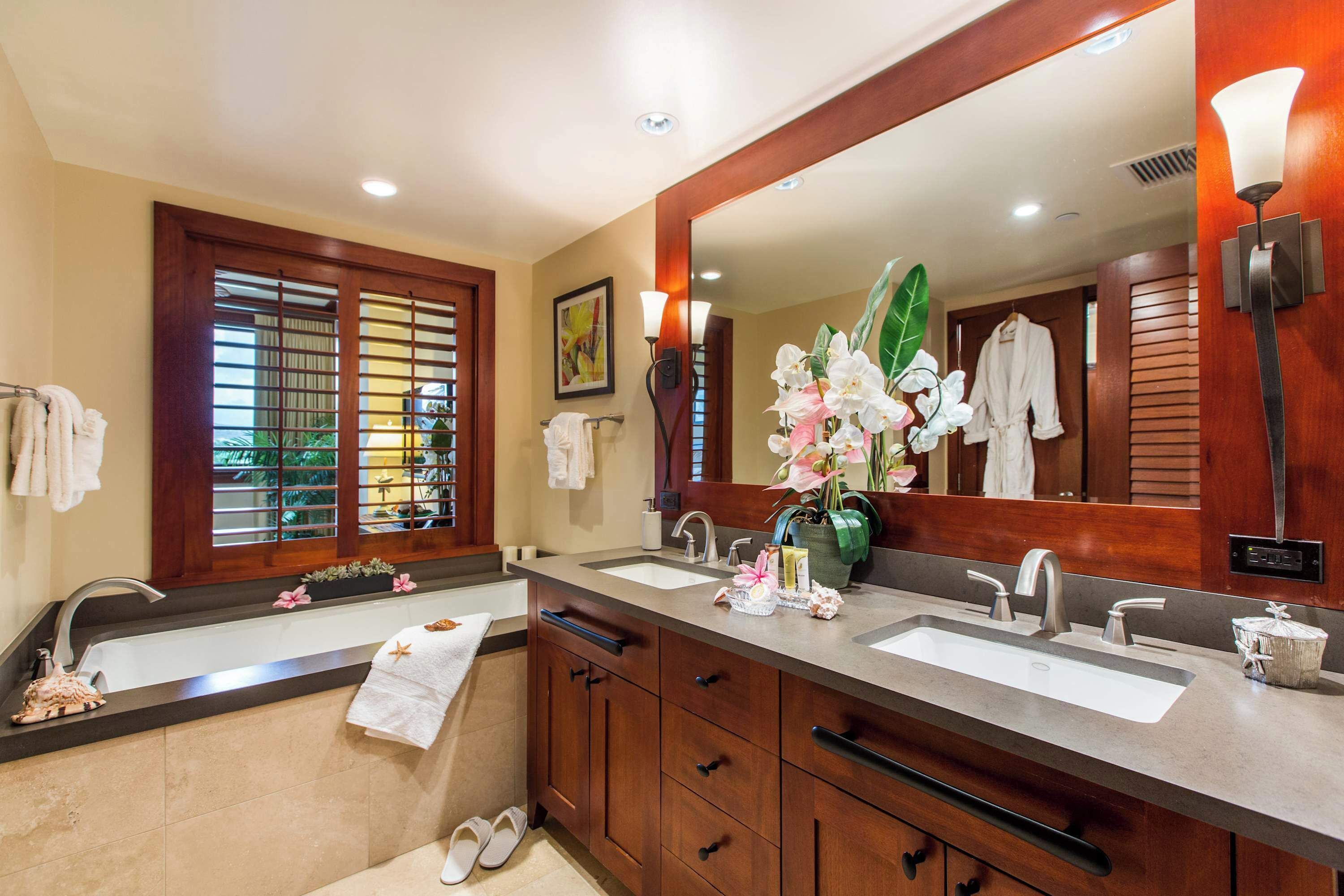 A modern bathroom with a large mirror, two sinks, wooden cabinets, and a window with blinds.