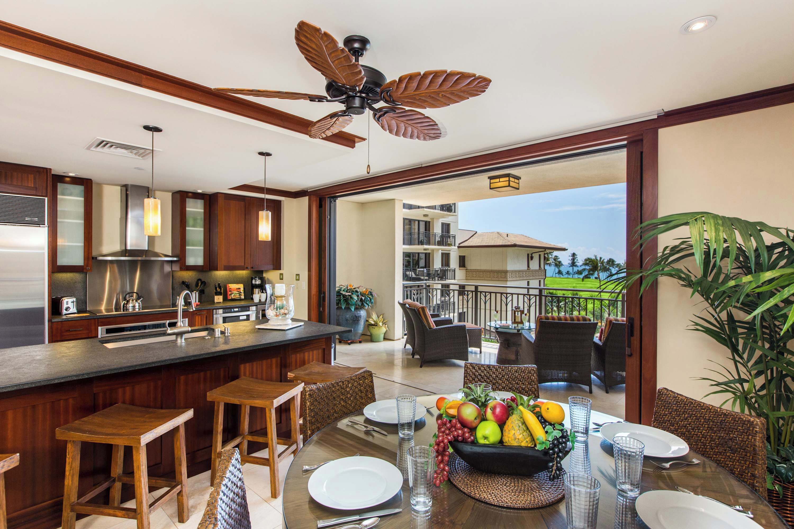 Cocina moderna con armarios de madera, una isla grande y una zona de comedor con sillas de mimbre y un frutero.