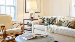 A cozy living room with a wicker chair, a white sofa with tree-patterned pillows, a round coffee table with a magazine, and a lamp on a side table. - Historic Nantucket Home | Hot Tub & Downtown Steps (Nantucket)