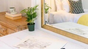 A bedroom with a large mirror, a bedside lamp, a bed with pillows, a dresser, and a desk with papers and a plant. - Historic Nantucket Home | Hot Tub & Downtown Steps (Nantucket)
