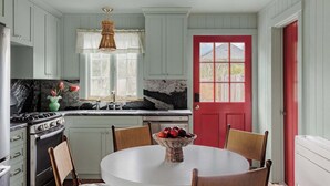 Une cuisine avec des meubles blancs, une porte rouge, une cuisinière à gaz et une table à manger avec des chaises.
