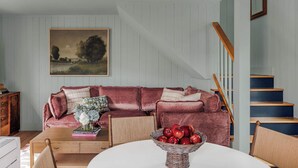 Un salon chaleureux avec un canapé en velours rouge, une table blanche sur laquelle se trouve un panier de pommes, et un escalier aux marches bleues.