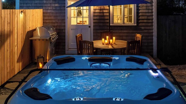 Un jacuzzi situé devant une maison dotée d’une terrasse en bois, d’un barbecue et d’une table décorée de bougies.