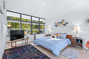 A modern bedroom with a large window, a flat-screen TV, a bed with blue bedding, a colorful rug, and a nightstand with a lamp. - Mid Century Modern Weekly Winter Rates! (Tampa)