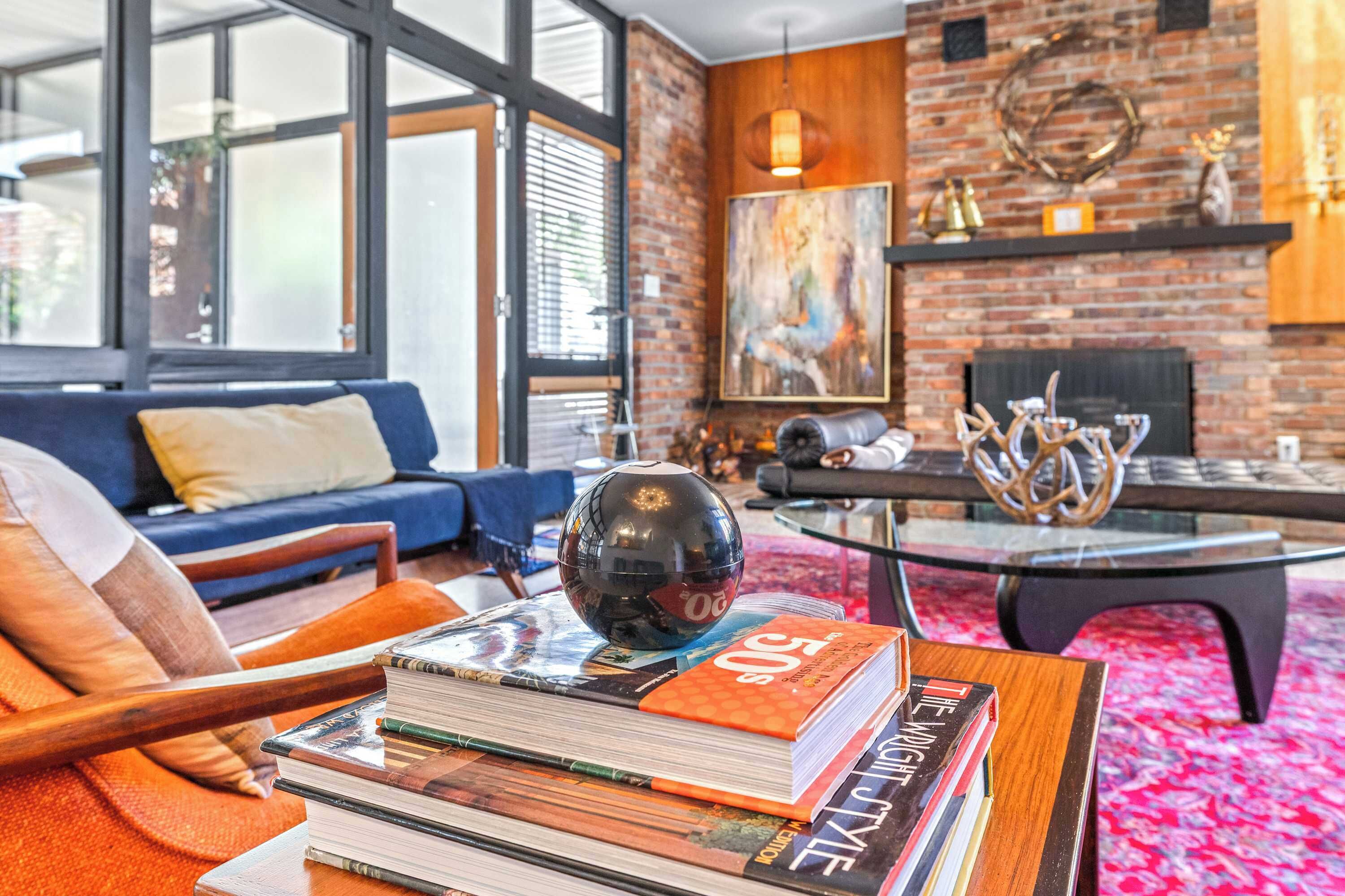 A modern living room with a sofa, a coffee table with books, a fireplace, and a large window.