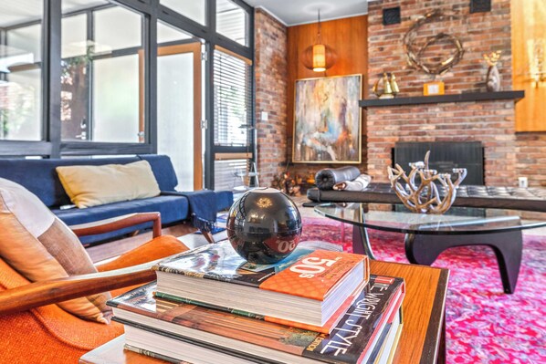 A modern living room with a sofa, a coffee table with books, a fireplace, and a large window. - Mid Century Modern Weekly Winter Rates! (Tampa)
