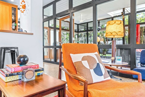 A modern living room with a large window, a wooden chair, a table with books and a radio, and a lamp. - Mid Century Modern Weekly Winter Rates! (Tampa)