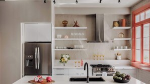 Una cucina moderna con frigorifero in acciaio inox, mobili bianchi, isola centrale con lavello e finestra con cornice rossa.