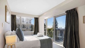 Una camera d'albergo con un letto, un comodino con una lampada, un'ampia finestra con tende e vista su edifici e alberi.