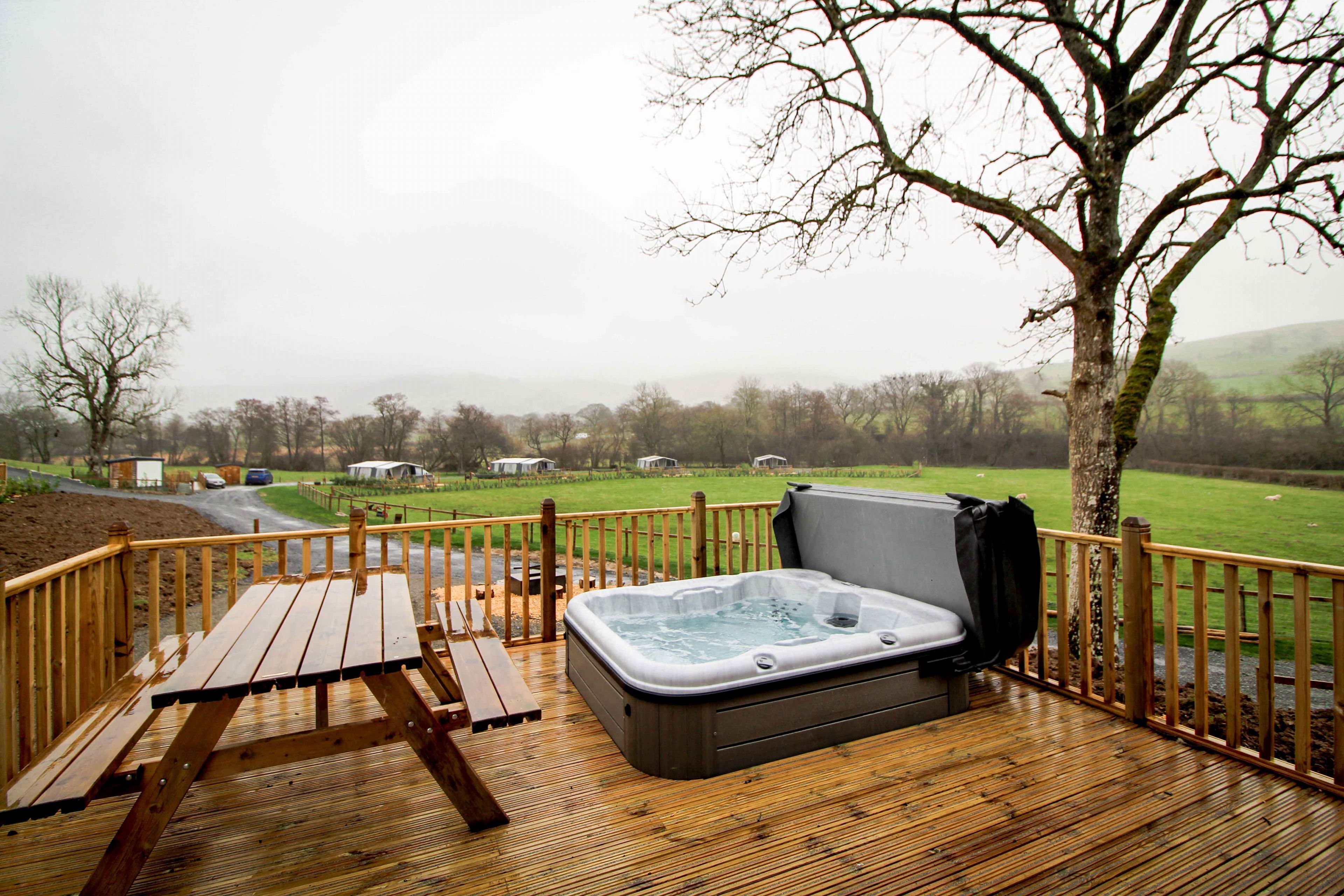 Eine Holzterrasse mit Whirlpool, Picknicktisch und Blick auf eine grüne Landschaft mit Bäumen und einem Zaun.