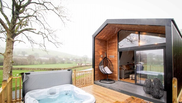 Un área moderna de hidromasaje al aire libre con terraza de madera, una silla colgante y una puerta de cristal que da a un espacio interior acogedor.