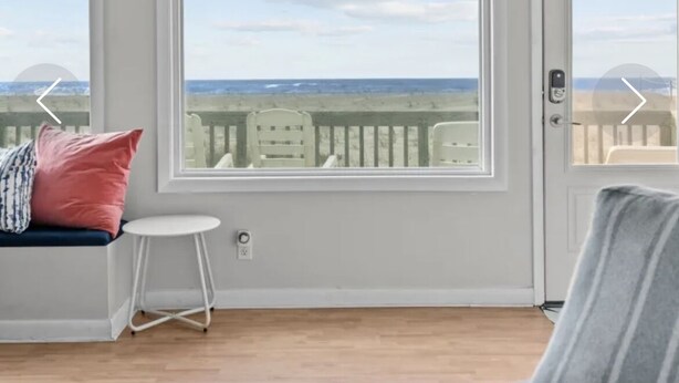 Una habitación con un amplio ventanal que ofrece vista a la playa y al océano. Hay un ventilador de techo, un banco blanco con cojines y una pequeña mesa blanca.