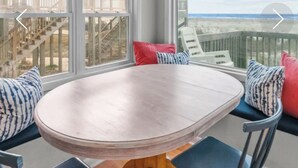 Una habitación iluminada por el sol, con una mesa redonda de madera y sillas, ventanas amplias que ofrecen vistas a una playa y una escalera.