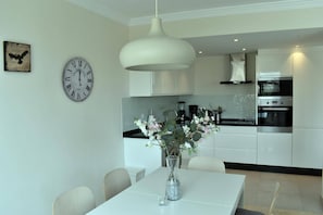 Een moderne keuken met witte kasten, een roestvrijstalen oven en een koffiezetapparaat. Er is een eettafel met stoelen en een vaas met bloemen. Op de muur staat een klok.