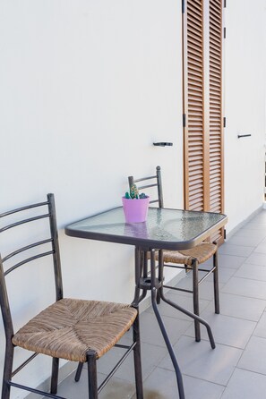 Una mesa con cubierta de vidrio, dos sillas de mimbre, una maceta morada y contra una pared blanca, persianas de madera.