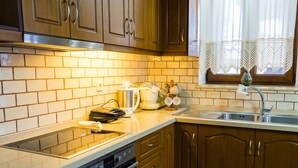 A kitchen with wooden cabinets, a stainless steel sink, and a window with lace curtains. - Thòlos Home (Αρχοντική)