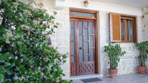A stone building with a wooden door and window, potted plants, and a tiled floor. - Thòlos Home (Αρχοντική)