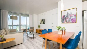 A modern living room with a sofa, coffee table, dining table, chairs, and a cityscape painting. - Infinite View| Balcony & Pool+ Gym+ Near Beach (Hallandale Beach)