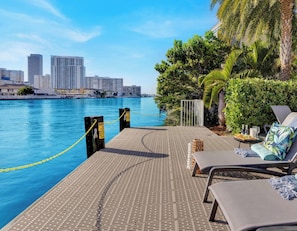 A waterfront pool area with lounge chairs, a palm tree, and a city skyline in the background.