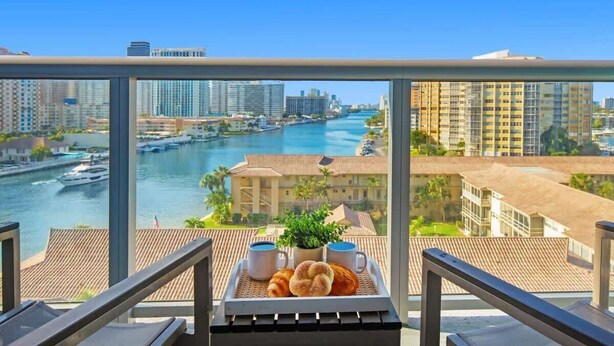 Un balcon avec un plateau de croissants, deux tasses et une plante, donnant sur une rivière bordée d’immeubles et de bateaux.