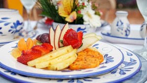 Uma travessa de frutas com morangos, maçãs e laranjas, acompanhada de um prato decorativo e um arranjo floral ao fundo.