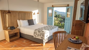 Quarto com cabeceira de madeira, cama com colcha estampada, telefone ao lado da cama e mesa de jantar com uma tigela de frutas e um jarro.