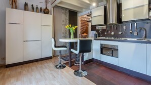 Een moderne keuken met witte kastjes, een roestvrijstalen afzuigkap en een ingebouwde oven. Er staan twee barkrukken bij een kleine tafel met een vaas bloemen.